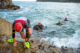 Three swimrunners in the sea, watching a fourth competitor scrambling out over a rocky outcrop.