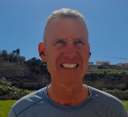A man wearing Nank blue tooth ear buds viewed from the front, with a blue sky in the background.
