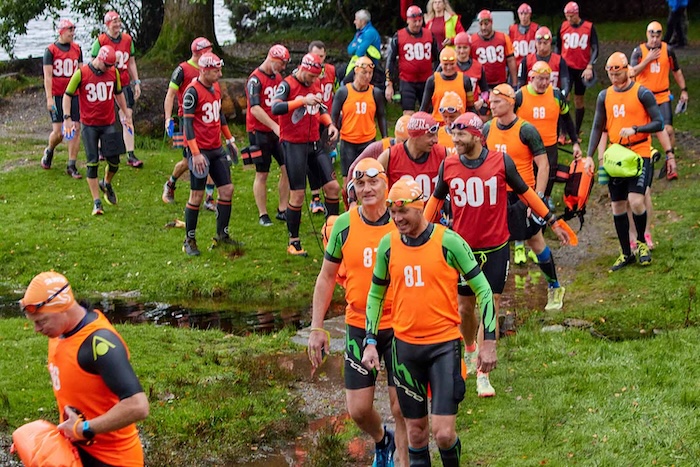 A group of men in wetsuits and colored tops walk down a grassy hill at a Gritty Rascals Events swimrun start