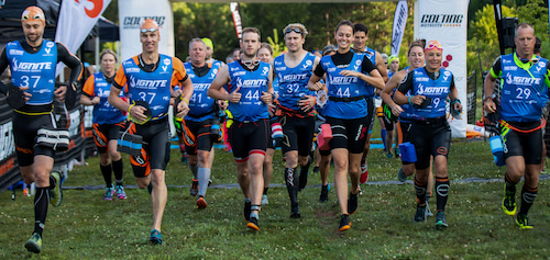 A group of swimrunners wearing blue tops and swim caps running across grass at the start of an IGNITE swimrun competition. A group of swimrunners wearing blue tops and swim caps running across grass at the start of an IGNITE swimrun competition.