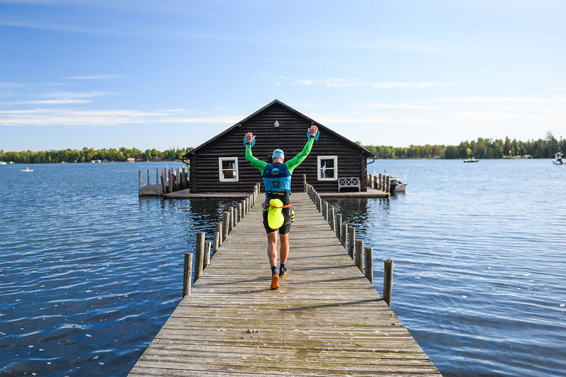 A man with a yellow float tied to his waist and his arms held up walking down a jetty towards a boat shed, with blue sky in the background. A man with a yellow float tied to his waist and his arms held up walking down a jetty towards a boat shed, with blue sky in the background.