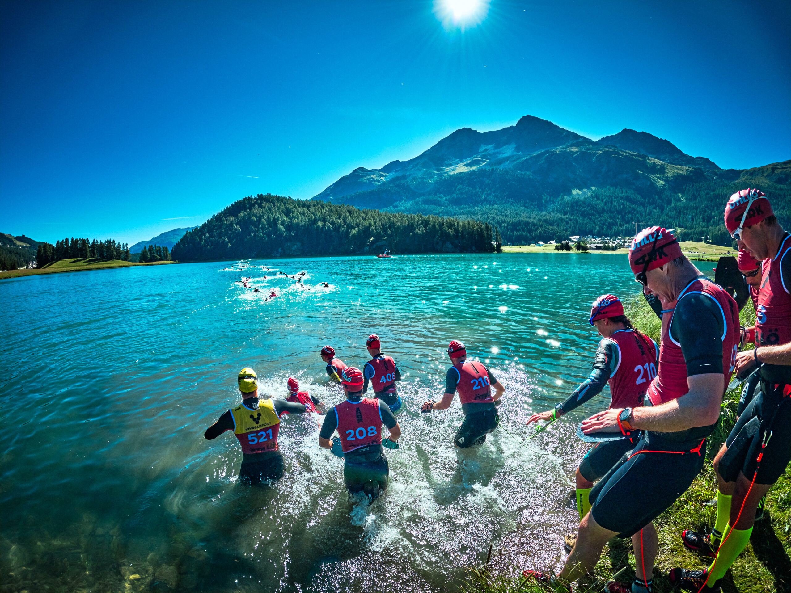 Otillo Engadin swim entry with athletes wading into a lake against a bright blue sky Otillo Engadin swim entry with athletes wading into a lake against a bright blue sky