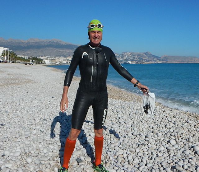 A man in a black wetsuit and orange socks with a green swim hat holding a shopping bag on a white stone beach with blue sky in the background