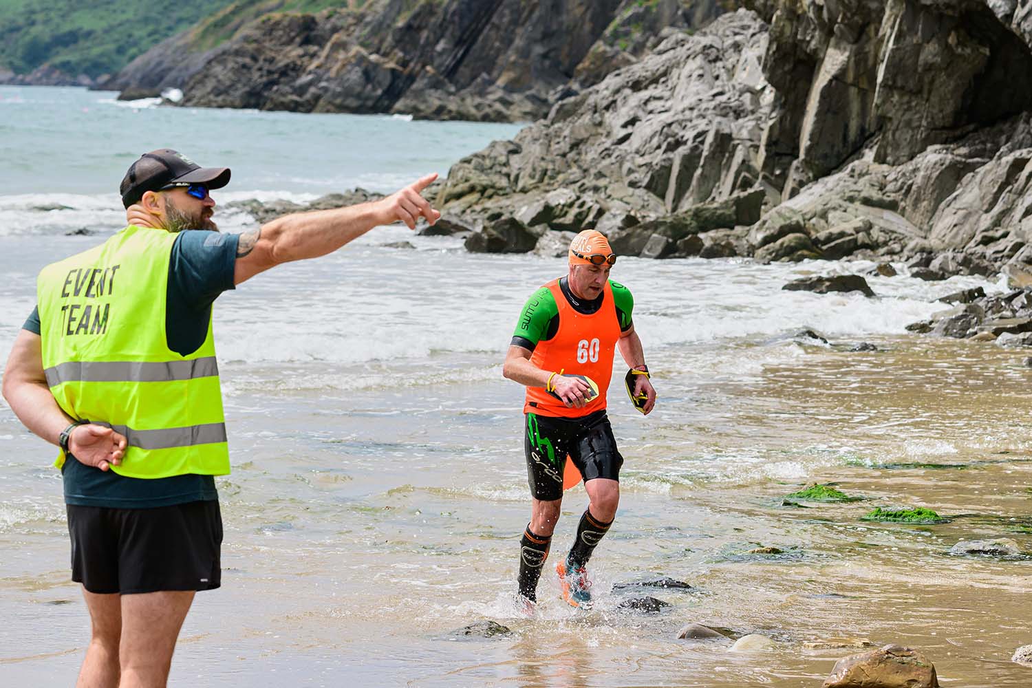 A man in an orange top running out of the sea, with another man in a yellow jacket pointing the way at the Swimrun Gower Beach Exit