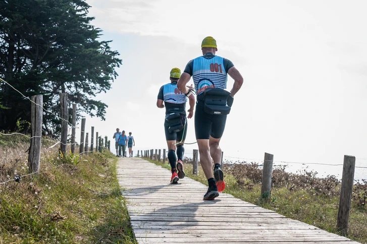 Swimrun competitors wearing blue tops running away from the camera along a wooden boardwalk on a beach at the  Swimrun Sucinio race. Swimrun competitors wearing blue tops running away from the camera along a wooden boardwalk on a beach at the  Swimrun Sucinio race.