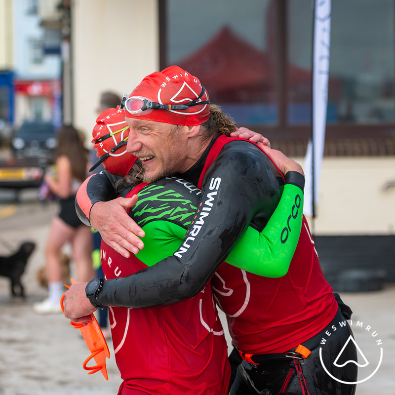 Two men in wetsuits and red tops hug at the Weswimrun event finish line. Two men in wetsuits and red tops hug at the Weswimrun event finish line.