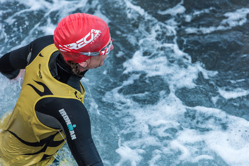 A swimrun racer wearing a yellow top and a red swim cap looking down at grey sea water