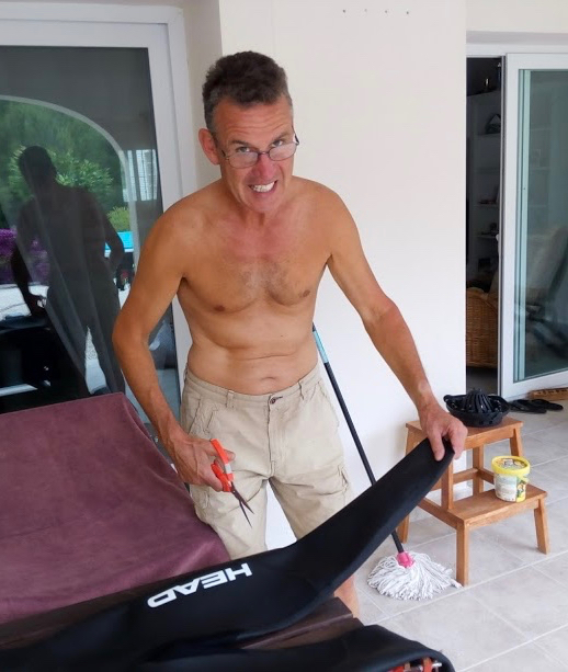 A man wearing shorts and reading glasses holding a wet suit sleeve and a pair of scissors, looking at the camera.
