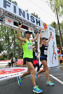 A man and woman cross the finish line at the Honolulu Marathon, holding their hands up.