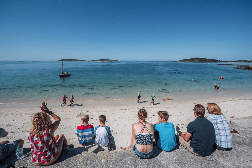 A small group of people sitting on a sand bank by blue water, watching the competitors in a swimrun race at the second swim leg at the Isles of Scilly, UK.