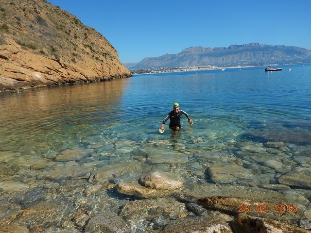 A man in a wetsuit and green cap scrambling over rocks out of the sea, from clear water
