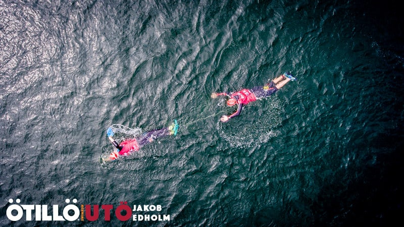 Two swimrunners viewed from a drone above a black lake, with the tether visible in the water.