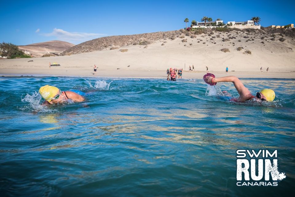 Two swimrunners wearing yellow caps swimming in blue sea with a sandy beach in the background, at the swimrun canary islands event. Two swimrunners wearing yellow caps swimming in blue sea with a sandy beach in the background, at the swimrun canary islands event.