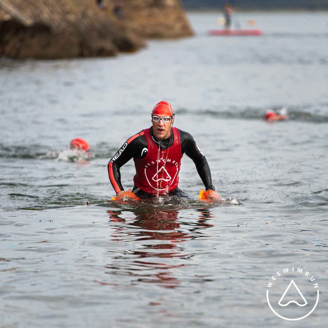 A man in a wetsuit and red top with swim goggles coming out of the water at the Weswimrun Dyfi X event A man in a wetsuit and red top with swim goggles coming out of the water at the Weswimrun Dyfi X event